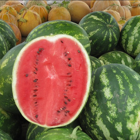 A pile of whole watermelons with one cut in half, showing the red flesh and black seeds. Other fruits are visible in the background.
